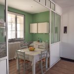 La photo montre une cuisine moderne et lumineuse. Les murs sont peints en vert clair, Sur la droite de la photo la cuisine est séparée des autres pièces par une baie vitrée. Une fenêtre donnant sur l’extérieur est placée sur le mur de gauche avec des rideaux blancs. sous la fenêtre se trouve un radiateur. sur le mur du fond se trouve un meuble bas et des éléments hauts blancs. Au centre de la cuisine se trouve une table avec une nappe cirée à fleurs et quatre chaises vertes en paille. Une corbeille de fruits est positionnée au centre de la table. La pièce est lumineuse.